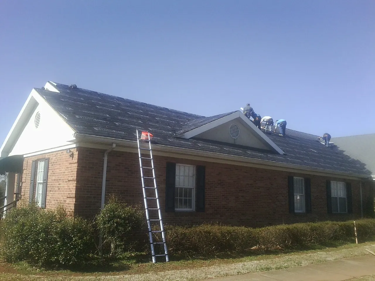 Crew tearing shingles off a brick ranch roof with a ladder set — Emergency Roof Repair work by our crew in Madill