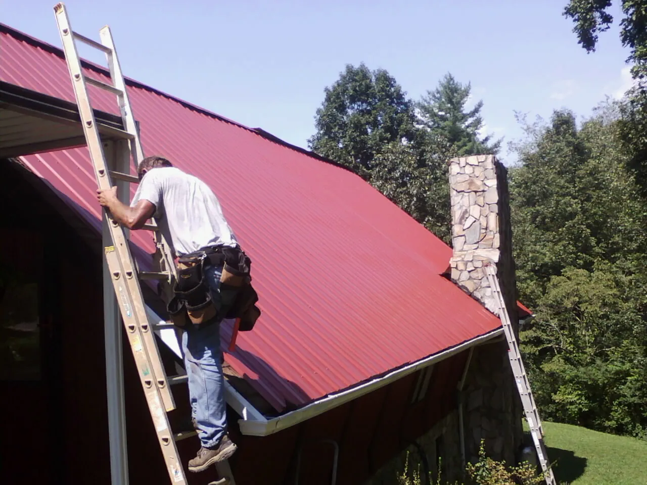 Roofer on a ladder beside a red metal roof and stone chimney — Soffit & Fascia Repair by Roofing Brothers in Madill